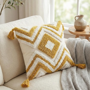 Könnte beinhalten: Quadratisches Zierkissen mit cremefarbenem Hintergrund und einem geometrischen Muster in Weiß und Senfgelb. Das Kissen hat Quasten an jeder Ecke und liegt auf einem cremefarbenen Sofa.