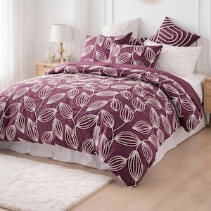 Puede incluir: Una cama con un edredón de color ciruela y fundas de almohada a juego, todas adornadas con un estampado de hojas blancas. La cama está hecha con sábanas y almohadas blancas. Una alfombra blanca está sobre el suelo de madera frente a la cama.