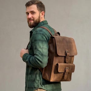 May include: A brown leather backpack with a flap closure and two front pockets. The backpack has adjustable straps and metal buckles. The leather has a distressed look. The backpack is worn on the back.