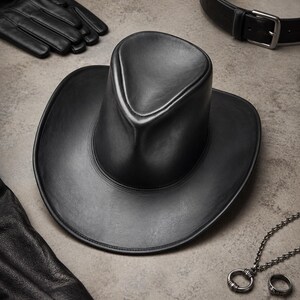 Puede incluir: Un sombrero de vaquero de cuero negro con una corona redondeada y ala ancha. El sombrero est&aacute; rodeado de guantes de cuero negro, un cintur&oacute;n negro con hebilla plateada y un collar plateado con colgante de anillo y un anillo a juego.