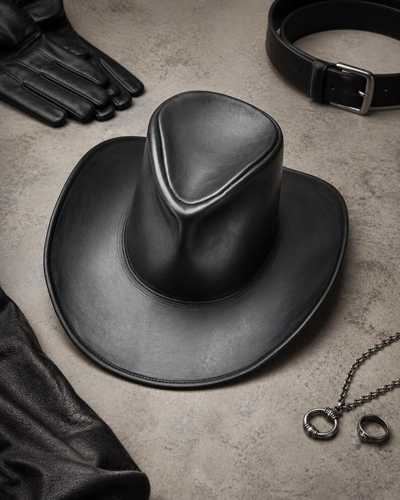 Puede incluir: Un sombrero de vaquero de cuero negro con una corona redondeada y ala ancha. El sombrero est&aacute; rodeado de guantes de cuero negro, un cintur&oacute;n negro con hebilla plateada y un collar plateado con colgante de anillo y un anillo a juego.