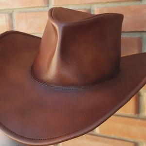 Puede incluir: Un sombrero de vaquero de cuero marr&oacute;n con ala ancha y copa redondeada. El sombrero es de un color marr&oacute;n rico y c&aacute;lido y parece estar hecho de cuero de alta calidad. El ala del sombrero es curvada y la copa tiene una ligera hendidura.