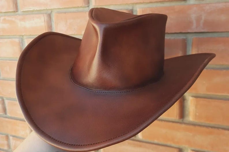 Puede incluir: Un sombrero de vaquero de cuero marr&oacute;n con ala ancha y copa redondeada. El sombrero es de un color marr&oacute;n rico y c&aacute;lido y parece estar hecho de cuero de alta calidad. El ala del sombrero es curvada y la copa tiene una ligera hendidura.