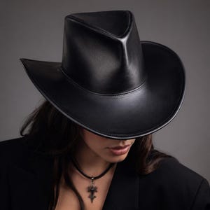 Puede incluir: Un sombrero de vaquero de cuero negro con una corona estructurada y ala ancha. El sombrero se lleva con una blazer negro y un collar con colgante. El dise&ntilde;o del sombrero es elegante y moderno.