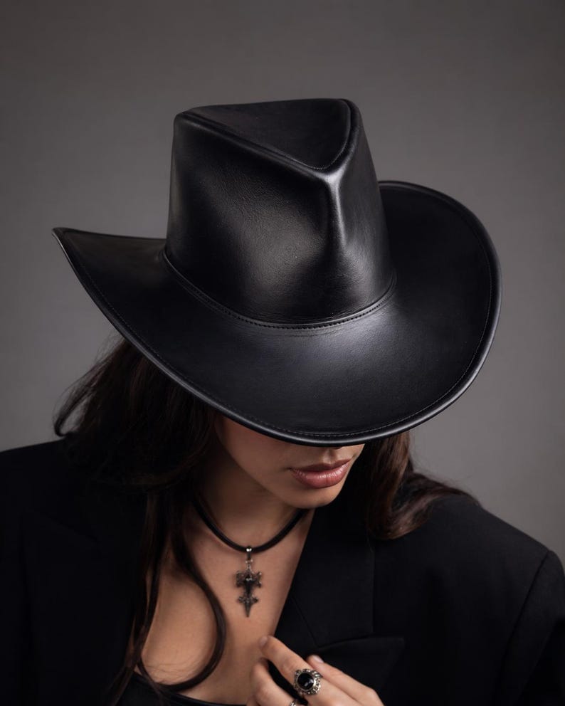 Puede incluir: Un sombrero de vaquero de cuero negro con una corona estructurada y ala ancha. El sombrero se lleva con una blazer negro y un collar con colgante. El dise&ntilde;o del sombrero es elegante y moderno.