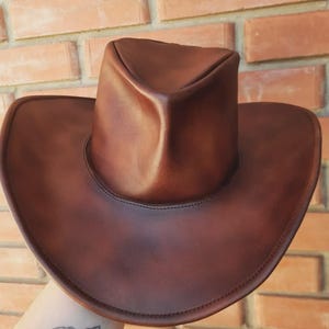 Puede incluir: Un sombrero de vaquero de cuero marr&oacute;n con ala ancha y copa redondeada. El sombrero es de un color marr&oacute;n oscuro intenso y parece estar hecho de cuero de alta calidad. El sombrero est&aacute; sostenido por una mano.