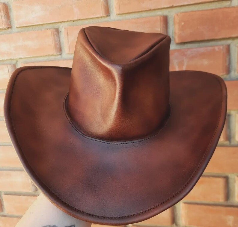 Puede incluir: Un sombrero de vaquero de cuero marr&oacute;n con ala ancha y copa redondeada. El sombrero es de un color marr&oacute;n oscuro intenso y parece estar hecho de cuero de alta calidad. El sombrero est&aacute; sostenido por una mano.