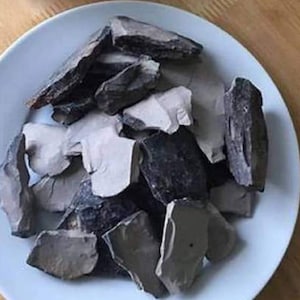 May include: A white plate holding a collection of grey, black, and off-white clay pieces. The clay fragments are of various sizes and shapes, some flat and others more irregular. The plate is white and rests on a wooden surface.