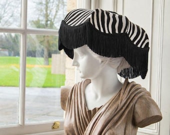May include: A lampshade featuring a black and white zebra stripe pattern with a scalloped bottom edge trimmed in long black fringe. The lampshade is positioned on a white classical bust, displaying the fabric pattern and the fringe detail.