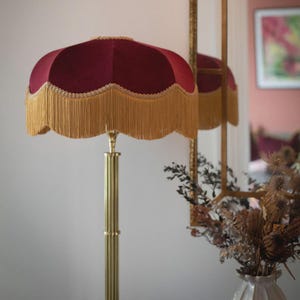 Puede incluir: Una lámpara de pie con una pantalla de terciopelo color burdeos profundo con borde festoneado, adornada con un ribete trenzado dorado y largos flecos dorados. La lámpara tiene un soporte de metal de latón pulido con textura estriada.