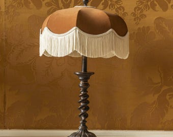May include: A table lamp featuring a brown, scalloped fabric shade with a long, cream-colored fringe trim. The lamp base is made of dark, weathered metal with a spiraling, twisted central column and a wide, ornate base with four small feet.