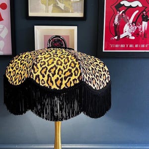 Può includere: Lampada decorativa con paralume con stampa leopardata. Il paralume ha un bordo smerlato con frange nere. La lampada ha una base color oro ed è posta contro un muro blu scuro con opere d'arte incorniciate.