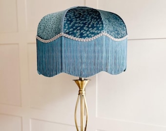Puede incluir: Una lámpara de pie con una pantalla de terciopelo azul cerceta con un patrón animal texturizado. La pantalla está adornada con un borde festoneado, un adorno plateado decorativo y largos flecos a juego. Se asienta sobre un soporte dorado.