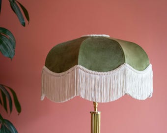 May include: A scalloped lampshade covered in olive green velvet fabric, featuring a decorative cream-colored braided trim along the edge and a long, straight cream-colored fringe hanging from the bottom, mounted on a fluted brass lamp stand.