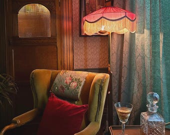May include: An interior scene featuring a velvet armchair, a fringed lamp, and a decanter. The armchair has a floral cushion and a red cushion. A glass of liquid sits on a small table next to the decanter.
