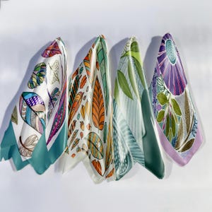 Puede incluir: Cuatro pañuelos de seda con coloridos diseños botánicos. Cada pañuelo presenta un patrón diferente, incluyendo plumas, hojas y motivos florales. Los pañuelos tienen bordes turquesa, verdes y lavanda y se exhiben sobre un fondo blanco.