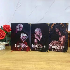 May include: Three hardcover books titled "Manacled" by SenLinYu. The book covers feature illustrations of figures in dark settings. Volume One shows a person holding a mask, Volume Two depicts a couple kissing, and Volume Three shows a couple embracing.