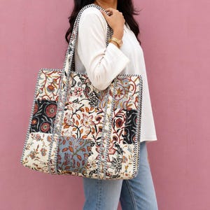 May include: A large rectangular tote bag featuring a patchwork design of various floral patterns in shades of cream, black, red, and blue. The bag has two shoulder straps with a black and white striped trim that matches the edges of the bag.