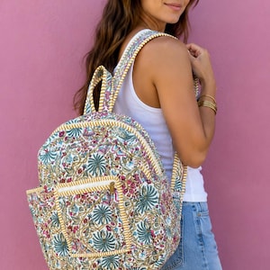 May include: A patterned backpack featuring a floral design in white, pink, and blue. It has a yellow trim and a front pocket. The straps and handle match the pattern. The background is a vibrant pink.
