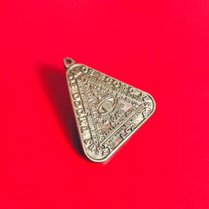 Pode incluir: Um pingente triangular prateado com gravuras detalhadas. O pingente apresenta um olho dentro de um triângulo, rodeado por texto. O fundo é vermelho sólido.