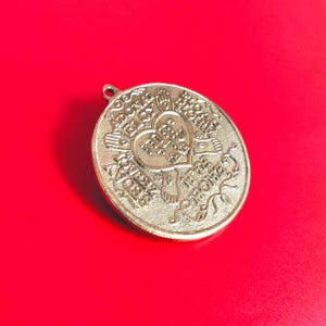 Puede incluir: Colgante dorado con un diseño en forma de corazón en el centro, rodeado de texto y elementos decorativos. El colgante tiene un lazo en la parte superior para colgar y está sobre un fondo rojo.
