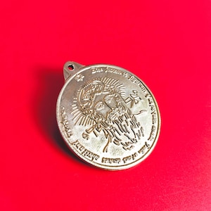 Puede incluir: Un colgante redondo de color plateado con un diseño en relieve que presenta una cara y texto. El colgante tiene un pequeño agujero en la parte superior para colgarlo. El fondo es rojo liso.
