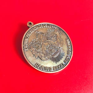 May include: A round, bronze-colored pendant with a loop at the top. The pendant features an embossed design with text and a central figure. The text includes "LA OMNI PRESENCIA DE DEUS" and "HARING BAKAL REX FERRUM".