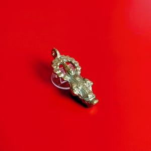 Puede incluir: Un colgante dorado que representa una figura religiosa con un halo, suspendido sobre un soporte de plástico transparente. El colgante presenta diseños detallados y se encuentra sobre un fondo rojo vibrante. El colgante es probablemente de metal.