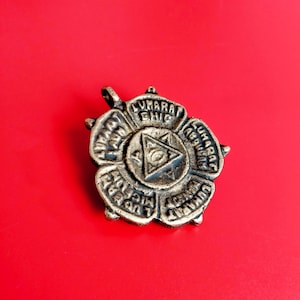 May include: A detailed, antique-style metal pendant with a floral design. The pendant features the text "LUMARA ABURAT" and "LUMARA MIC" around a central triangle with an eye symbol. The pendant is set against a red background.