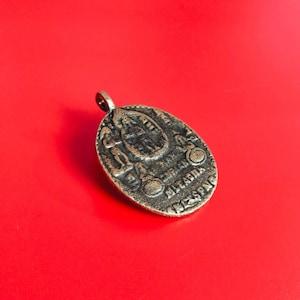 Könnte beinhalten: Ein antiker, bronzefarbener ovaler Anhänger mit aufwendigen geprägten Designs. Der Anhänger zeigt Figuren, Symbole und Text, mit einer Schlaufe oben für eine Kette. Der Hintergrund ist einfarbig rot.