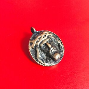 Pode incluir: Pingente oval de bronze com uma representação detalhada de uma figura religiosa com uma coroa de espinhos. O pingente tem uma argola na parte superior para uma corrente e está sobre um fundo vermelho vibrante.