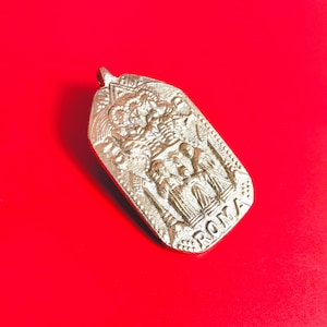 Pode incluir: Pingente prateado com uma cena religiosa em relevo e a palavra "ROMA" na parte inferior. O pingente tem formato retangular com uma argola na parte superior para uma corrente. Fundo vermelho.