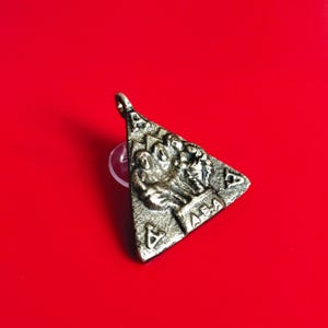 Puede incluir: Un colgante triangular plateado con un diseño en relieve detallado. El colgante presenta figuras y símbolos, incluido un triángulo dentro de un triángulo. El fondo es rojo liso.