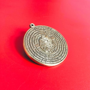 Puede incluir: Un colgante redondo de color bronce con un diseño y texto en relieve. La imagen central presenta una representación detallada de una figura con un halo. El anillo exterior contiene escritura. El colgante tiene un pequeño lazo en la parte superior para una cadena.