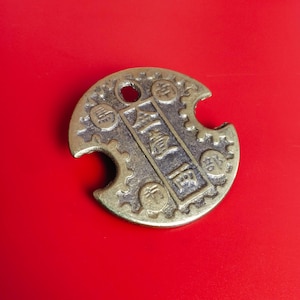 Op de afbeelding: Een bronzen, ronde munt met een gat aan de bovenkant en een hap uit de zijkant. De munt heeft Chinese karakters in het midden en een tandwielachtig ontwerp rond de rand. Op een helderrode achtergrond.