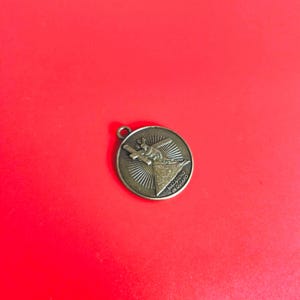 Puede incluir: Un colgante redondo de color bronce con un diseño religioso. El colgante presenta una imagen de una persona llevando una cruz, con las palabras "NAZARENO de QUIAPO" inscritas alrededor del borde. Tiene un pequeño lazo en la parte superior.