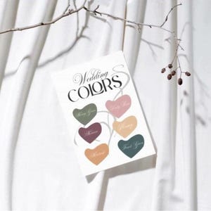 以下が含まれることがあります： 「Wedding COLORS」の文字と、緑、ピンク、オレンジなど様々な色のハート型のカラーサンプルが描かれた白いカード。白い背景に、枝から吊り下げられています。