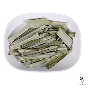 Op de afbeelding: Een witte, ovale kom gevuld met gedroogde, lichtgroene citroengrasbladeren. De bladeren zijn lang, dun en gelaagd, met een licht gekruld uiterlijk. De afbeelding is goed belicht en benadrukt de textuur en kleur van de kruiden.
