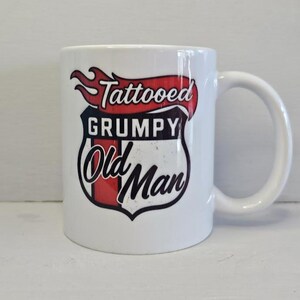 May include: White ceramic mug with a large handle. The mug features a graphic with the words "Tattooed Grumpy Old Man" in a shield design with red and black accents. The mug is on a white surface.