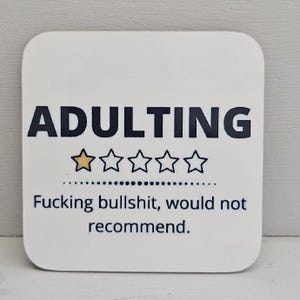 Op de afbeelding: Witte vierkante onderzetter met afgeronde hoeken. De tekst "ADULTING" is in het zwart gedrukt, boven een beoordeling van één gouden ster en vier lege sterren. Daaronder staat de tekst "Fucking bullshit, would not recommend."