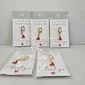 Könnte beinhalten: Mehrere kleine, goldfarbene Karabiner-Anhänger mit roten Herzanhängern werden auf weißen Karten präsentiert. Auf den Karten steht "A LITTLE HUG TO SAY A BIG" und "thank you" mit einem roten Herz. Die Anhänger sind in Klarsichtfolie verpackt.