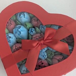 May include: A heart-shaped red gift box with a clear window lid, tied with a matching red satin ribbon. Inside the box are numerous small, sculpted flower-shaped items in shades of light blue, dusty rose, and muted green, arranged closely together.