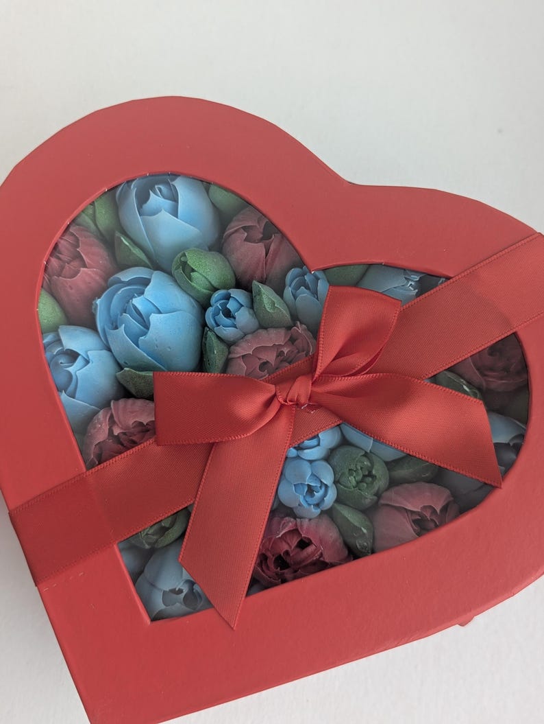 May include: A heart-shaped red gift box with a clear window lid, tied with a matching red satin ribbon. Inside the box are numerous small, sculpted flower-shaped items in shades of light blue, dusty rose, and muted green, arranged closely together.