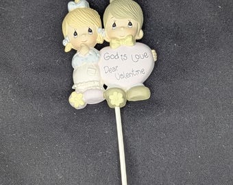 May include: A figurine of two children with blonde hair, holding a heart-shaped sign with the text "God is Love Dear Valentine." The children are dressed in pastel colours. The figurine is attached to a white stick.