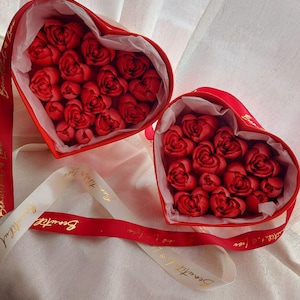 May include: Two heart-shaped red gift boxes filled with red rose-shaped items, lined with white tissue paper. A red ribbon with the gold text Beautiful Love is wrapped around the boxes, with additional white ribbon accents featuring the same text.