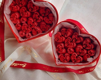 May include: Two heart-shaped red gift boxes filled with red rose-shaped items, lined with white tissue paper. A red ribbon with the gold text Beautiful Love is wrapped around the boxes, with additional white ribbon accents featuring the same text.
