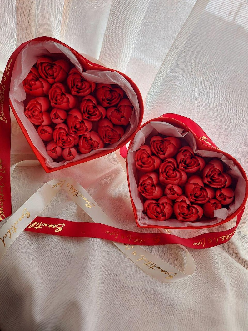 May include: Two heart-shaped red gift boxes filled with red rose-shaped items, lined with white tissue paper. A red ribbon with the gold text Beautiful Love is wrapped around the boxes, with additional white ribbon accents featuring the same text.