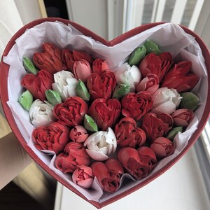 May include: A heart-shaped red box filled with a collection of red and white tulip-shaped confections and small green leaves. The arrangement is nestled inside white tissue paper, creating a dense display of floral-inspired treats.