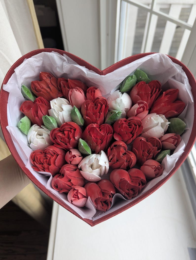May include: A heart-shaped red box filled with a collection of red and white tulip-shaped confections and small green leaves. The arrangement is nestled inside white tissue paper, creating a dense display of floral-inspired treats.