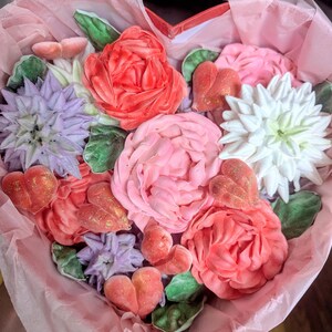May include: A heart-shaped box filled with decorative floral-shaped confections in shades of red, pink, white, and purple. The arrangement includes detailed flower blossoms, green leaves, and small heart-shaped pieces with a gold glitter finish.
